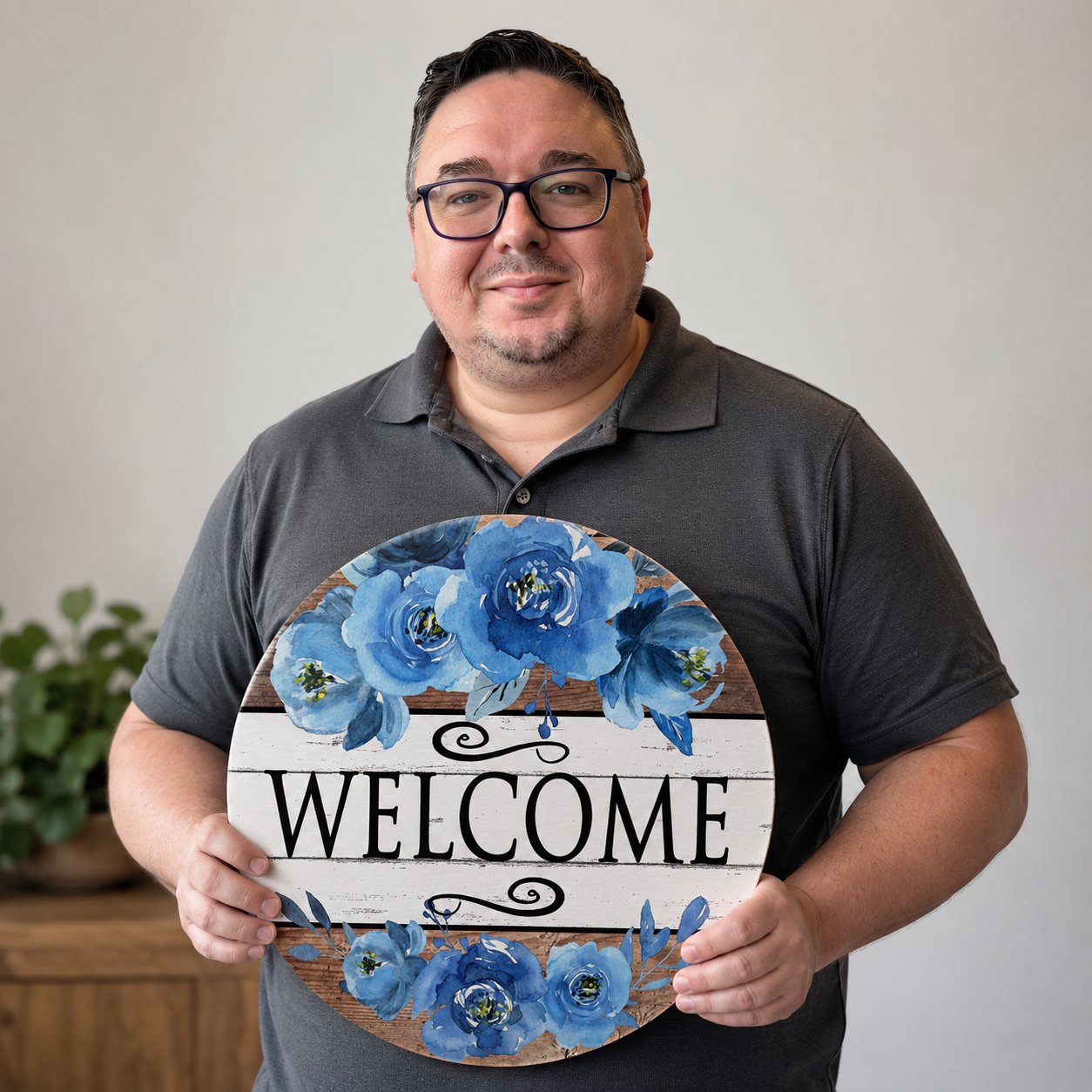 John Norrell, founder of The Crafty Tech Guy, smiling while holding a blue floral Welcome metal sign, showcasing the handcrafted home décor and personalized customer service behind the brand.