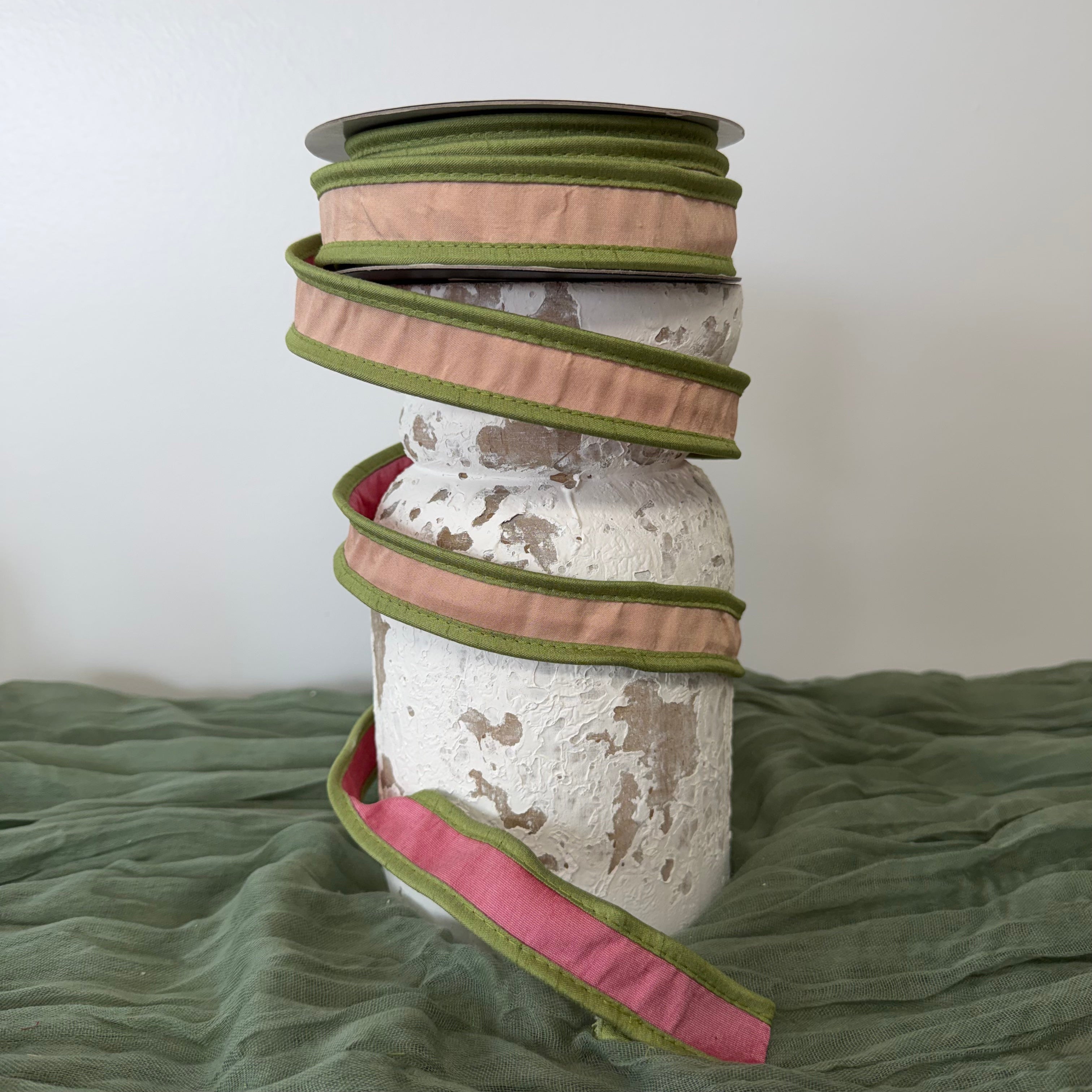 Roll of 1 inch pink double dupioni wired ribbon with green piping edges displayed on a decorative pedestal.