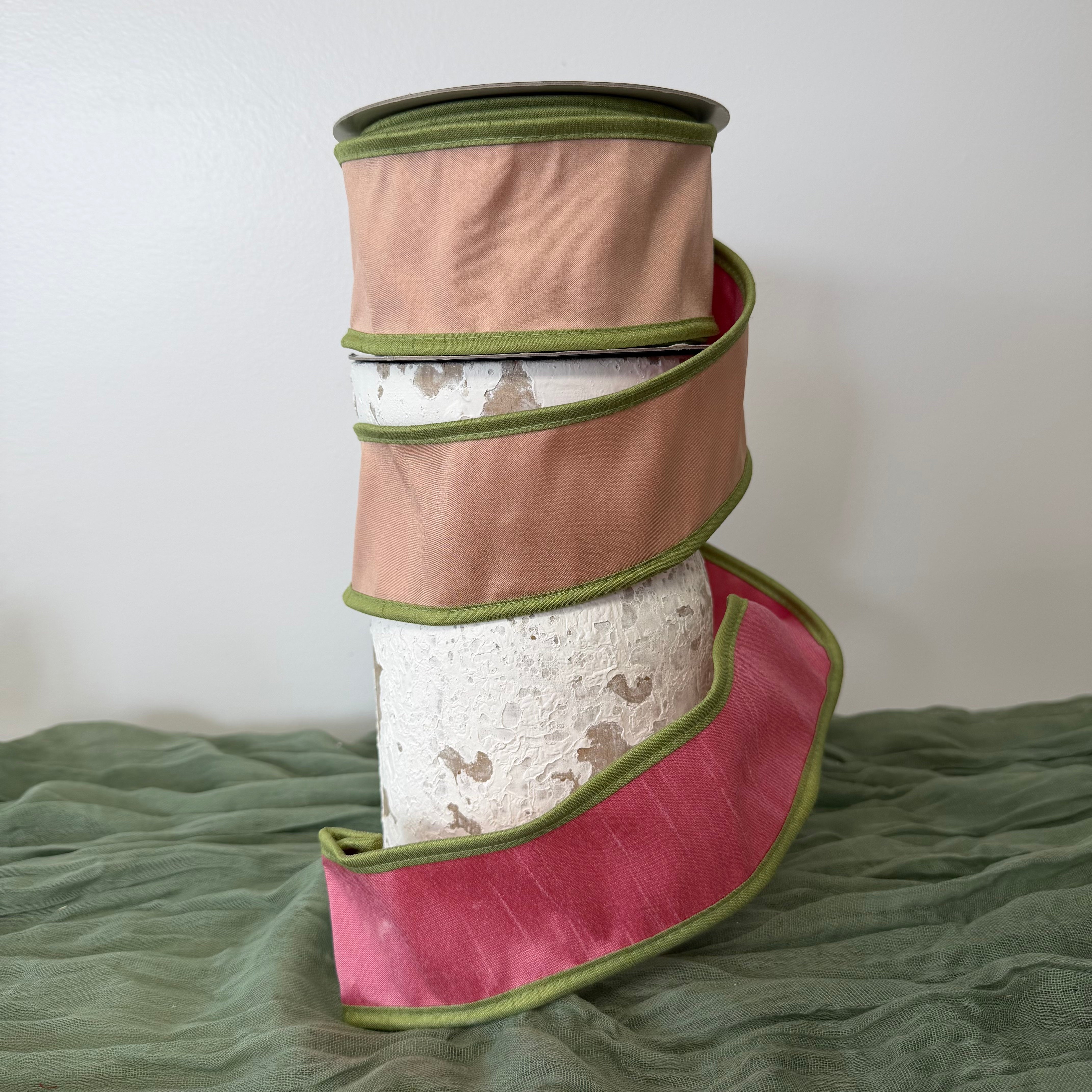Roll of 2.5 inch pink double dupioni wired ribbon with green piping edges displayed on a decorative pedestal.