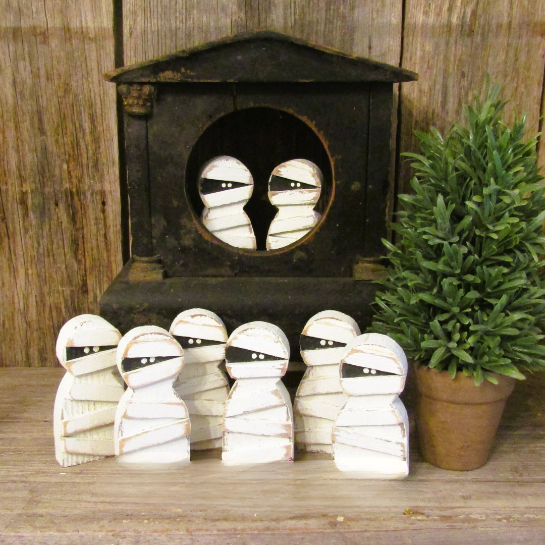 Group of small wood mummy Halloween decorations with painted bandages in front of a rustic black clock and greenery